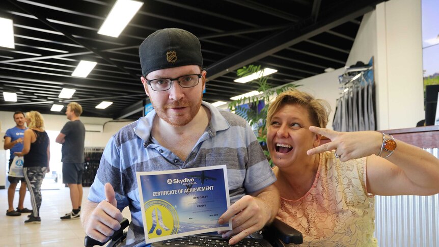Man in wheelchair holds a skydiving certificate of achievement with his thumbs up, next to a smiling woman making peace sign.