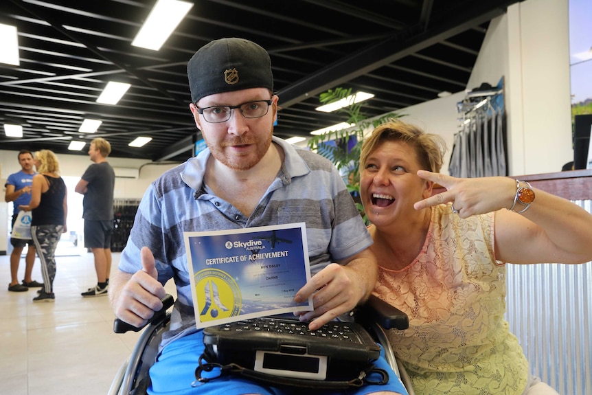 Man in wheelchair holds a skydiving certificate of achievement with his thumbs up, next to a smiling woman making peace sign.