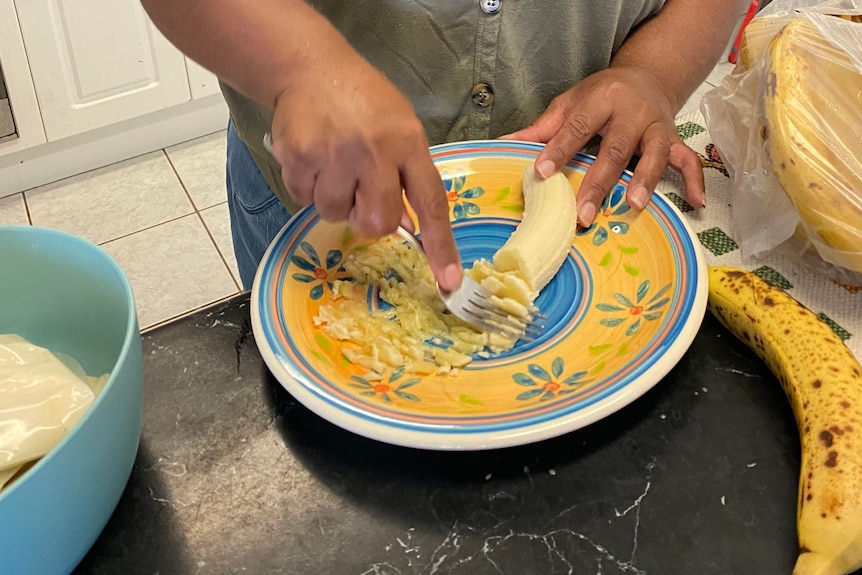 Woman mashing banana with fork onto plate. 