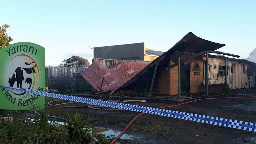 Partially-collapsed blacked building cordoned off by police tape. Smoke drifts from the site. Puddles and hoses surround it