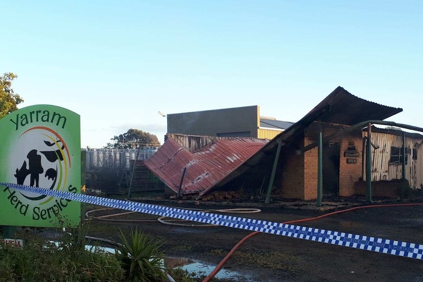 Partially-collapsed blacked building cordoned off by police tape. Smoke drifts from the site. Puddles and hoses surround it