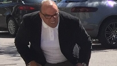 A bald man wearing a sports coat and white shirt squats behind a vehicle.