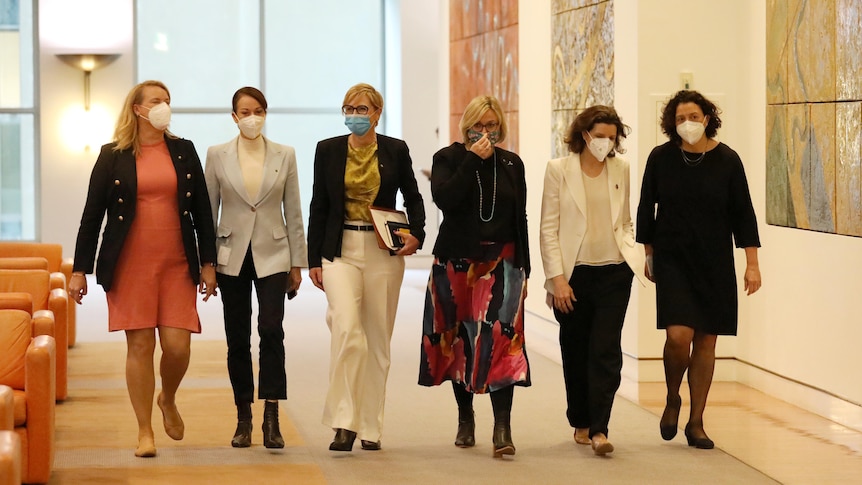 A row of women wearing masks walks towards the camera down a corridor.