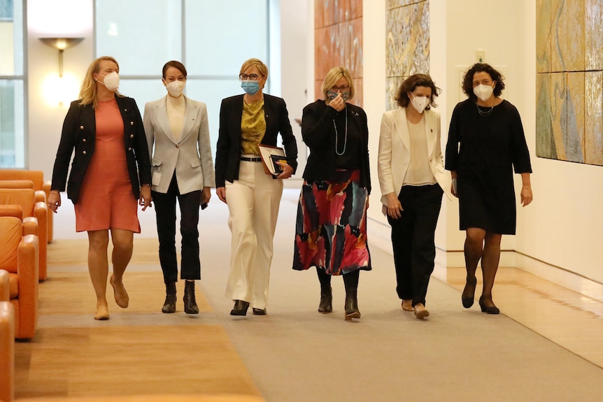 A row of women wearing masks walks towards the camera down a corridor.