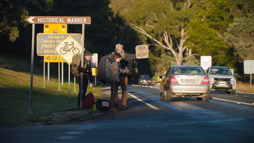 A woman, a man and a child with a sign saying 'India via Ballarat' standing beside a road.