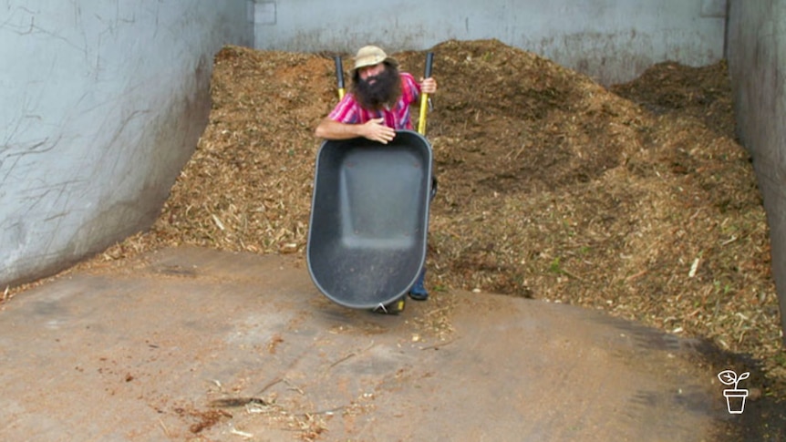 Man in hat standing in a bay holding mulch with tilted wheelbarrow in front of him