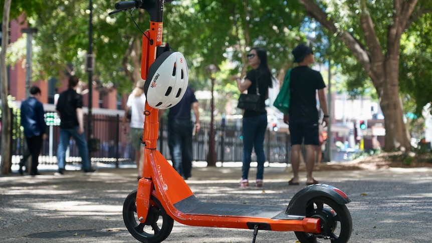 Orange e-scooter left parked on pathway with pedestrians walking in the background at South Brisbane.