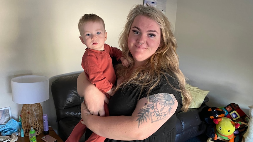 A smiling blonde woman with a large tattoo on her arm stands in her living room, holding her baby son.
