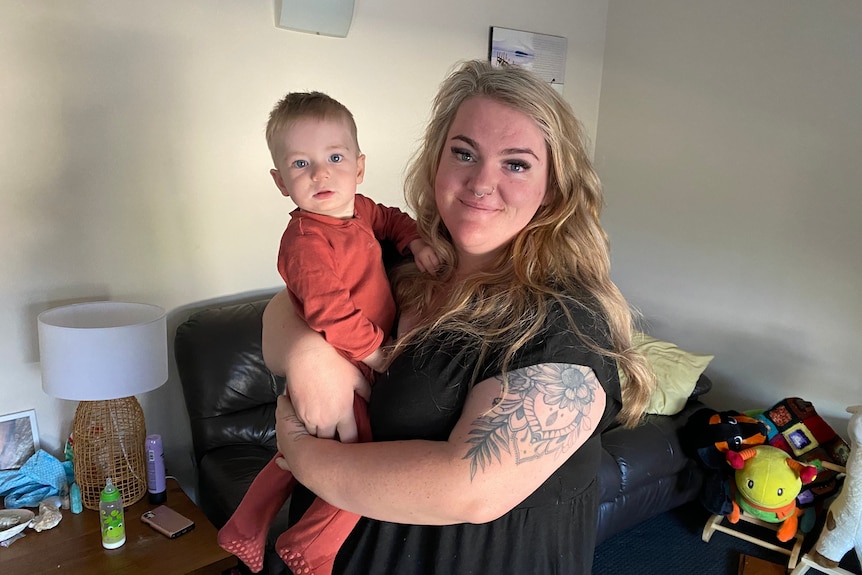 A smiling blonde woman with a large tattoo on her arm stands in her living room, holding her baby son.