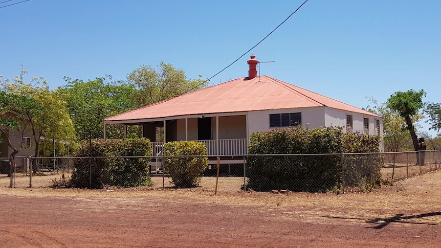 A cream weatherboard house with a red roof surrounded by shrubs and red dirt roads.