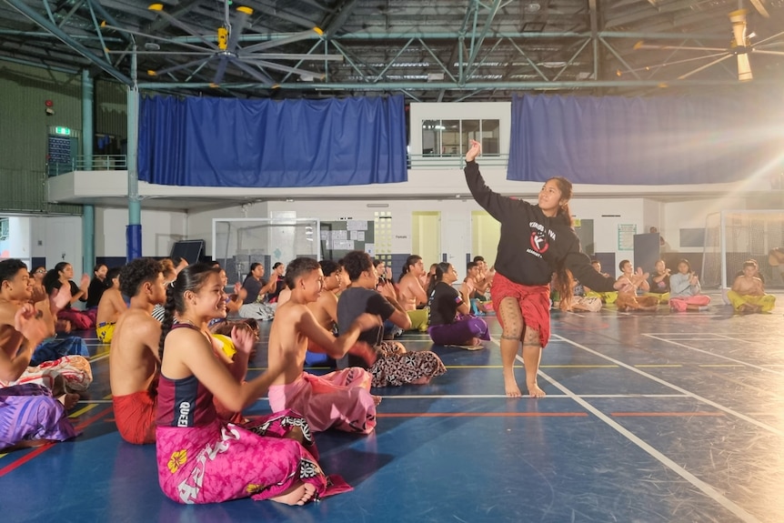 School aged teens sit around young woman dancing the siva. All students wear ie lava lava.