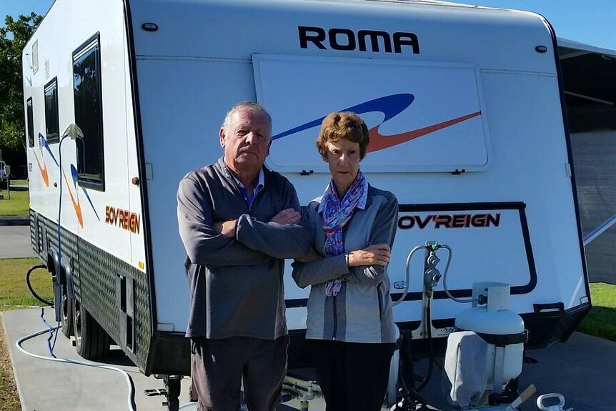 Man and woman stand in front of caravan with arms crossed.