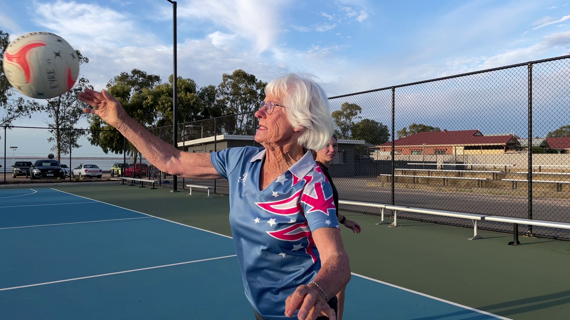 An older woman passing a netball on an outdoor netball court. Behind her the sky is blue. She has white hair.