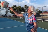An older woman passing a netball on an outdoor netball court. Behind her the sky is blue. She has white hair.
