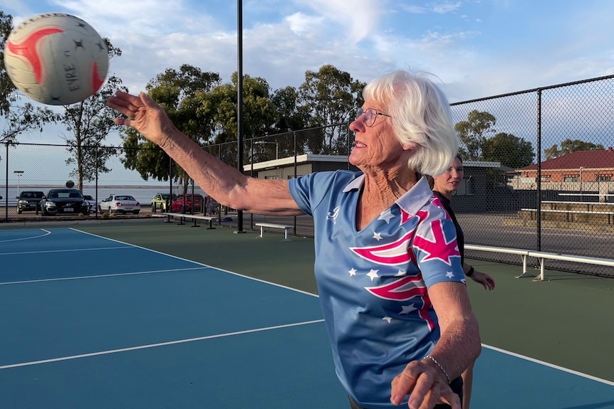 An older woman passing a netball on an outdoor netball court. Behind her the sky is blue. She has white hair.