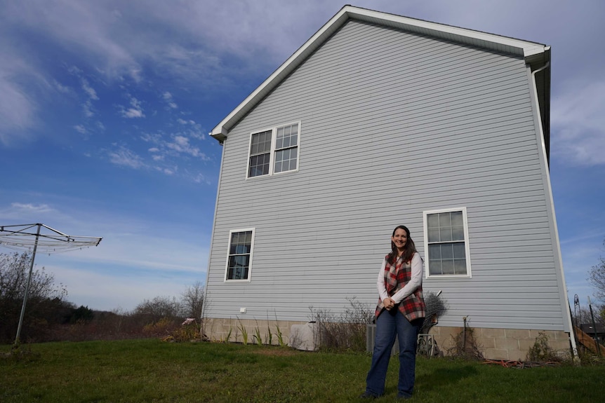 A woman in a tartan vest smile against the backdrop of a two-storey waterboard house