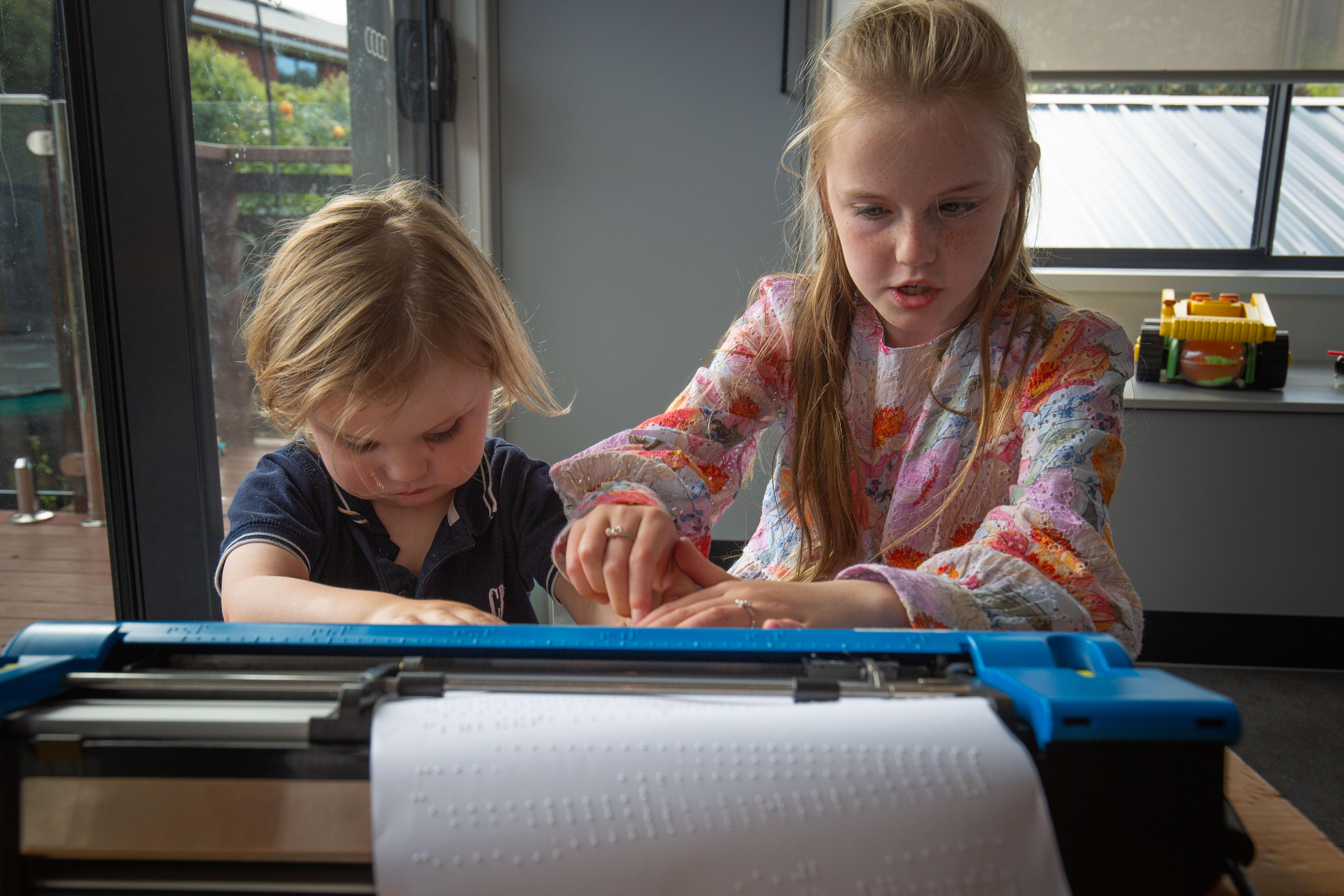 Una niña sostiene los dedos de su hermano menor para guiarlos hacia los botones de una máquina Braille. 