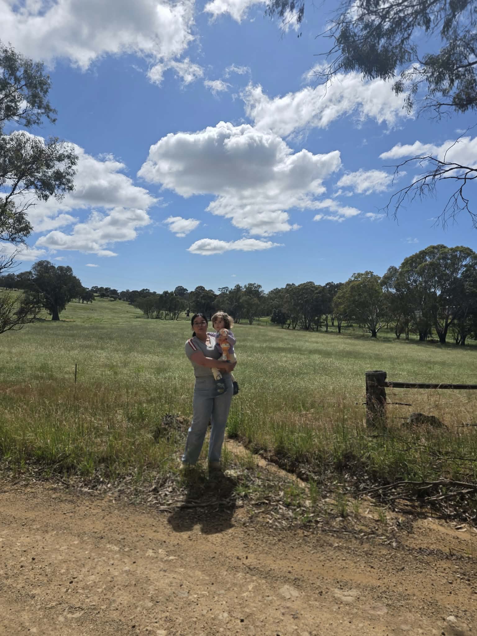 A woman in a grey top and jeans holding a young child in front of a large patch of open field.