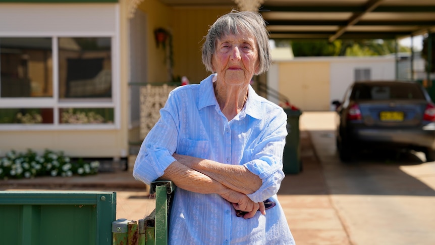 An elderly white woman in a blue shirt leaning on a green fence out the front of her Broken Hill home. 