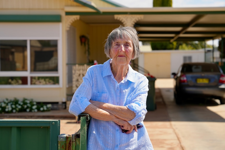 An elderly white woman in a blue shirt leaning on a green fence out the front of her Broken Hill home. 