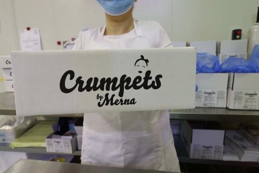 A woman in a white apron holding a box labelled Crumpets by Merna.