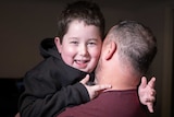 A young boy smiles and looks cheekily over the shoulder of his dad, who is carrying him.