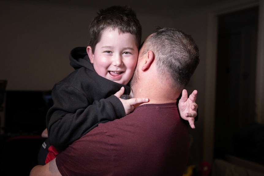 A young boy smiles and looks cheekily over the shoulder of his dad, who is carrying him.