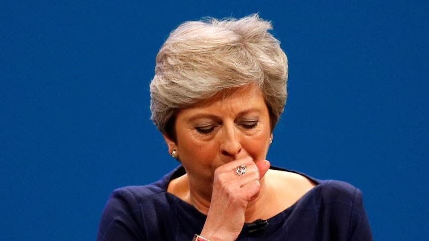 UK Prime Minister Theresa May in a blue dress, holding her fist to her mouth as she coughs during an address