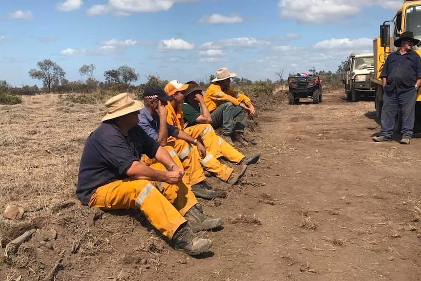 Men in yellow rural fire fighting gear and hats sit on the side of a road.