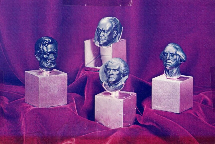 An old photo of four rocks carved into heads, propped up on blocks with a velvet/fabric looking background.