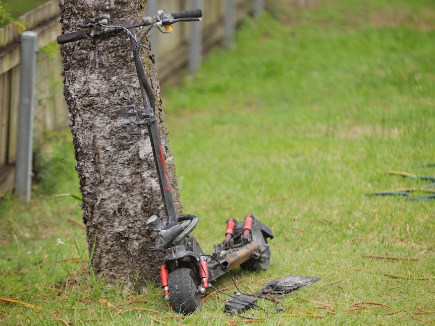 A damaged scooter that caught fire leans against a tree in a grassed yard