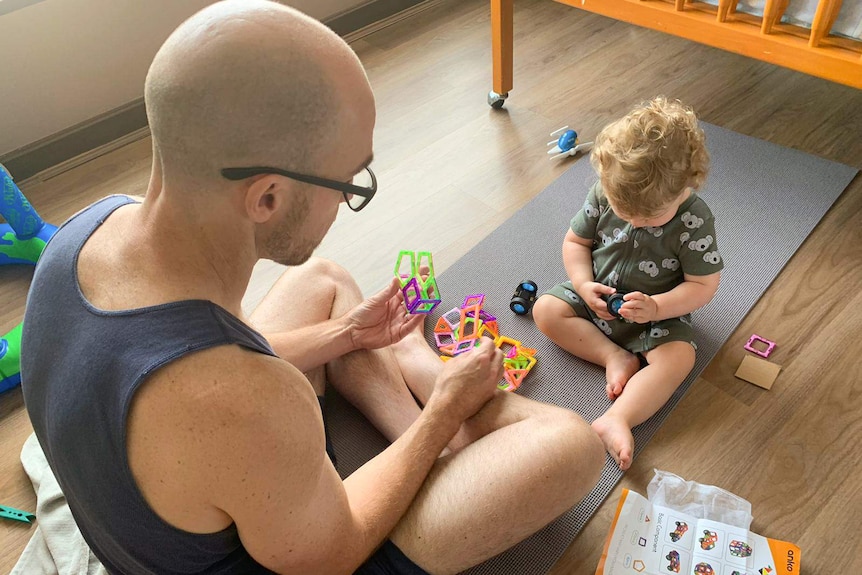 A man wearing glasses holds children's toys as he sits in front of a boy playing with a toy.