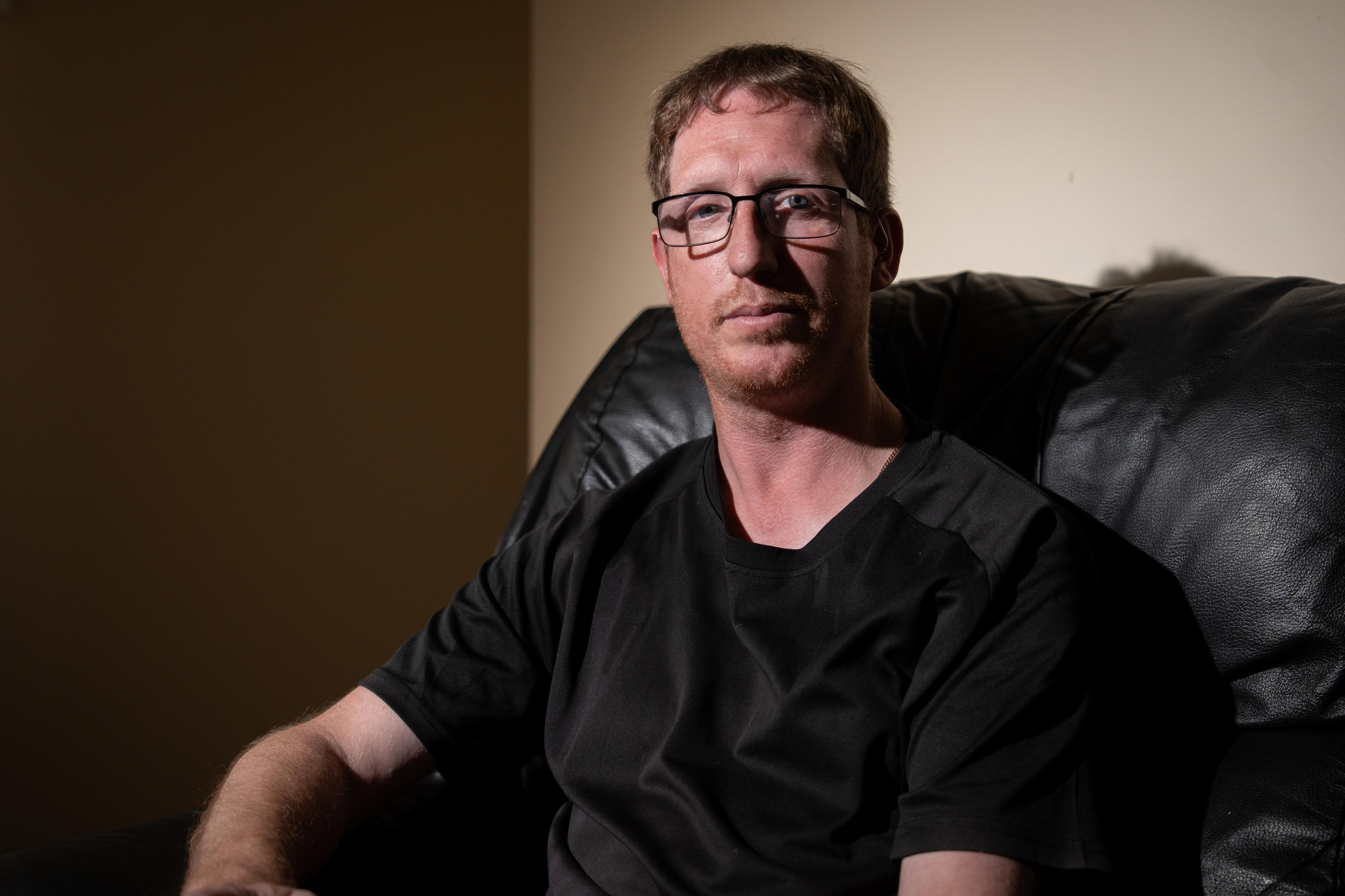 A man in glasses and a dark shirt sits on a leather couch.