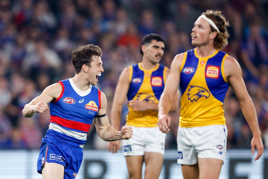 A grinning Western Bulldogs AFL player runs away from goal pumping his fists as a West Coast Eagles defender looks on.