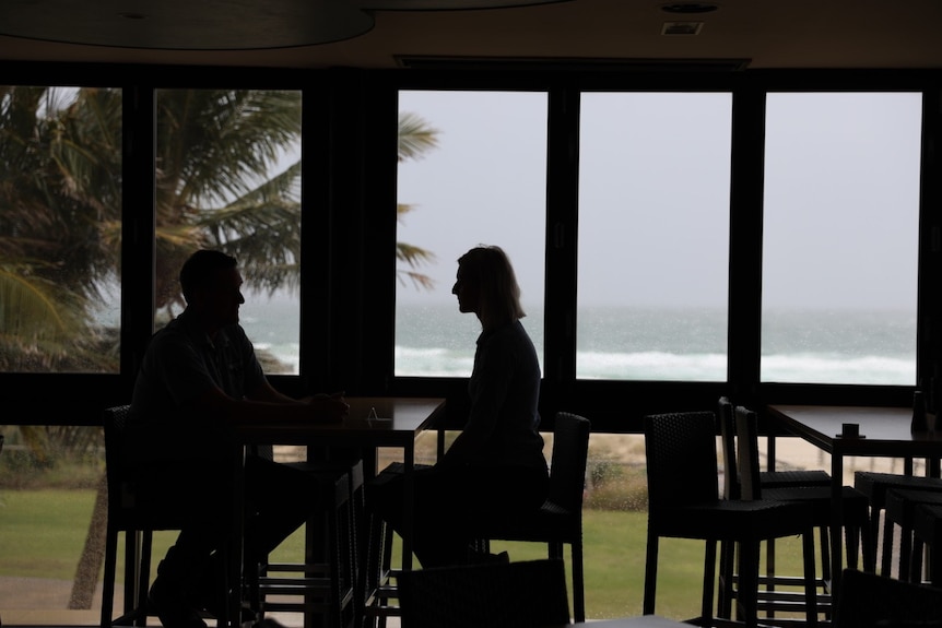 Two blacked-out figures sitting at a table talking against ocean through window