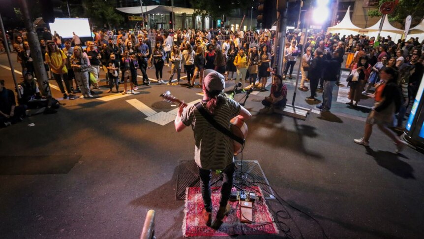 A crowd gathers around to watch a plan play the guitar while standing on a small mat. February 17, 2018.