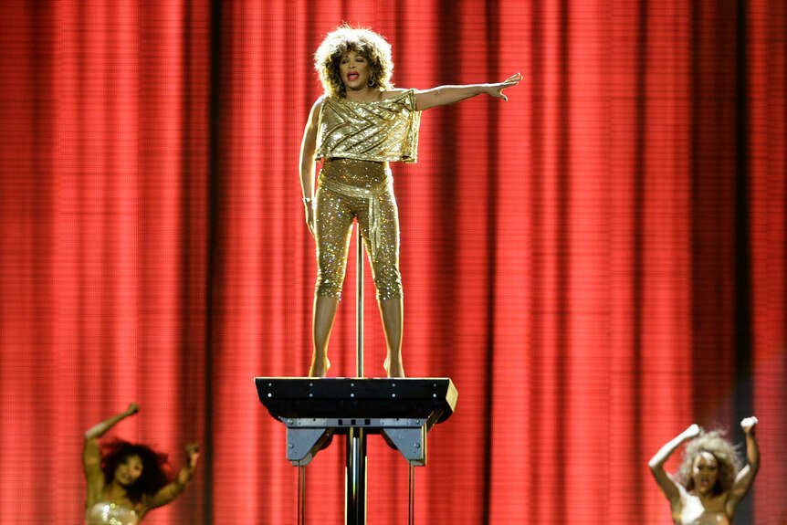 Tina Turner performing on a raised platform on stage. She's wearing a pair of sparkly gold pants and matching top. 
