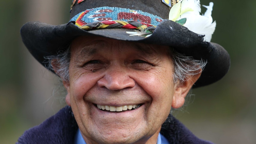 An Aboriginal man wearing a wide brim hat adorned with badges and features smiles to camera