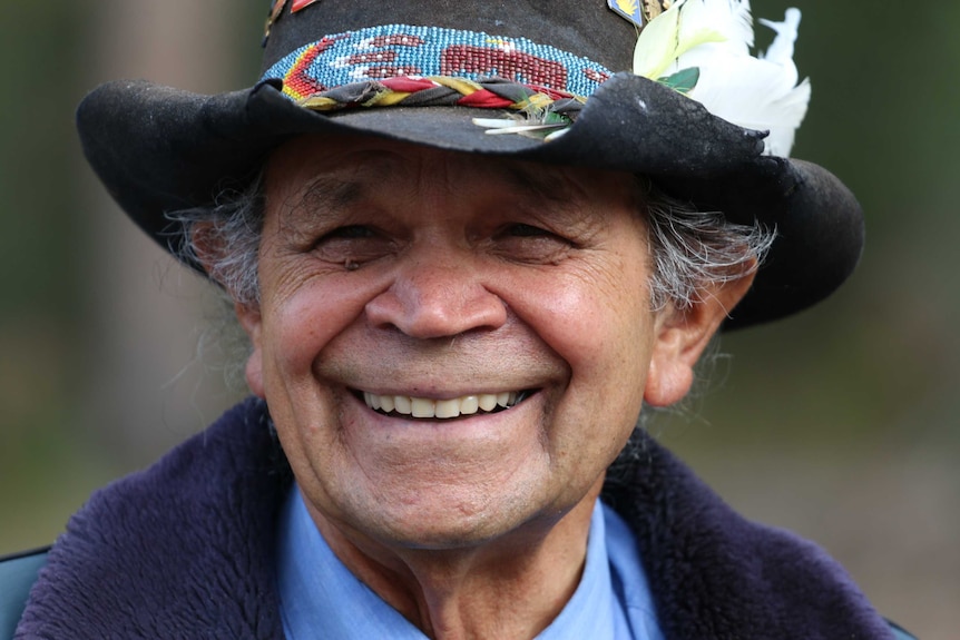 An Aboriginal man wearing a wide brim hat adorned with badges and features smiles to camera