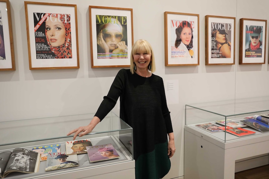 A distinguished woman leans against a glass case filled with magazine covers.