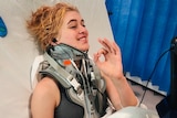 A teenager sits in a hospital bed with a neck brace on. 