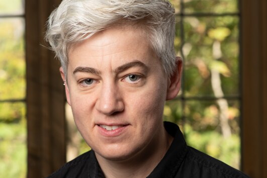 Non-binary person with short platinum hair open mouth smiles in front of window, inside