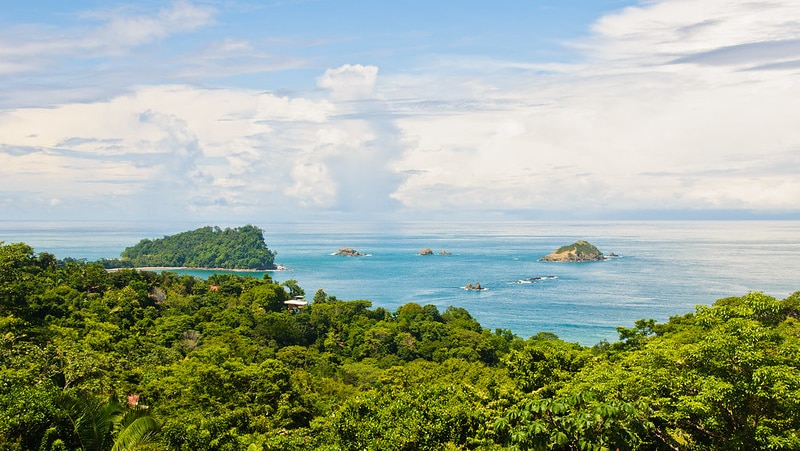 On a bright day, you look over a verdant tropical canopy to azure ocean waters with small islands in the distance.