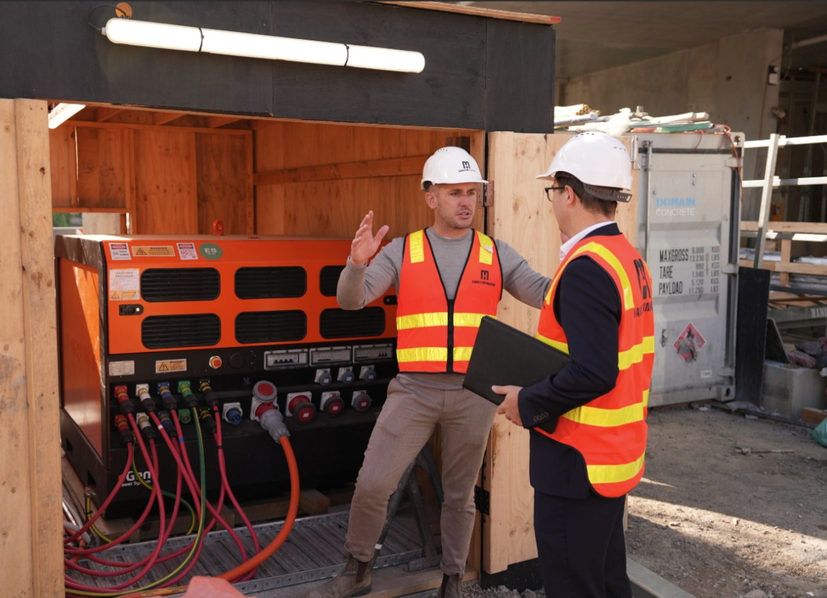 Two men in high-vis vests and helmets stand next to a large machine, talking to each other and gesturing.