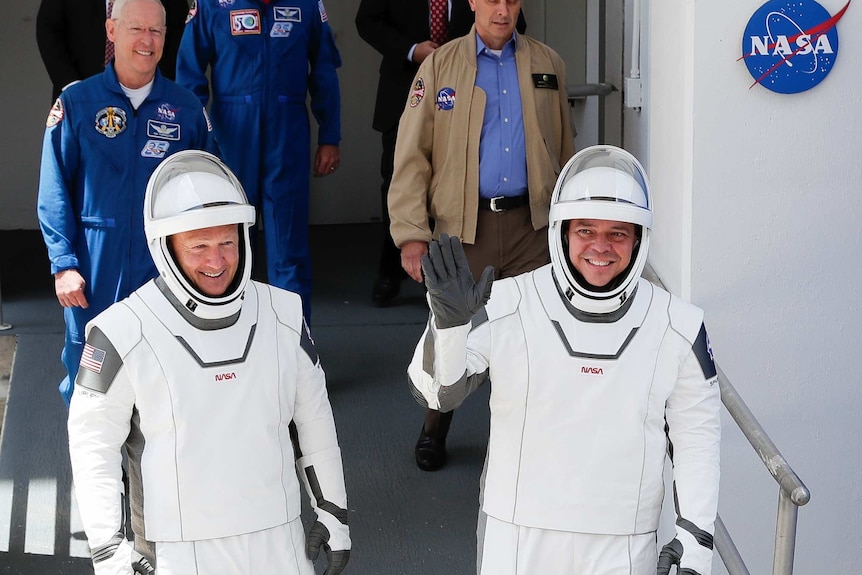 NASA astronauts walk in front of a white wall with a NASA sign wearing their spacesuits and waving.