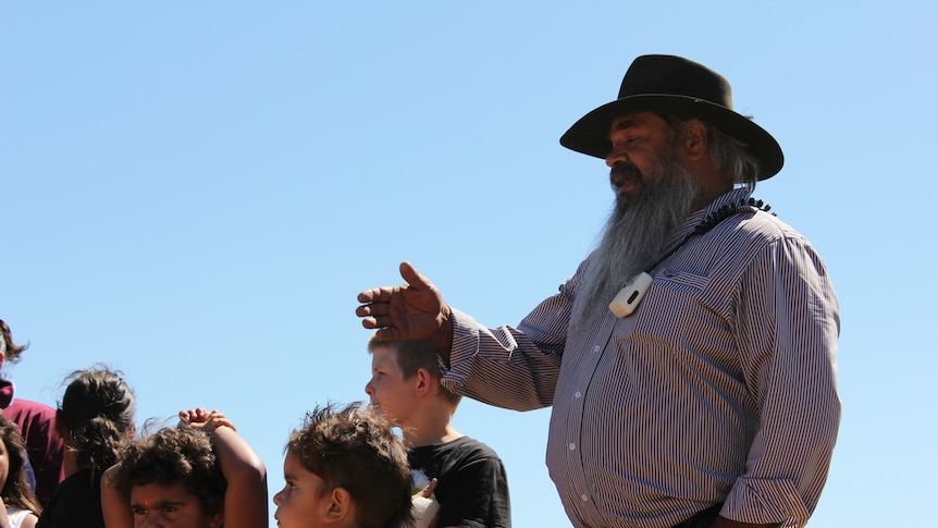 An Indigenous man with a beard and hat on the right, with children on the left.