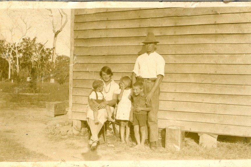 An old sepia picture of a man standing and a woman sitting with three children against a weatherboard house on stills.  