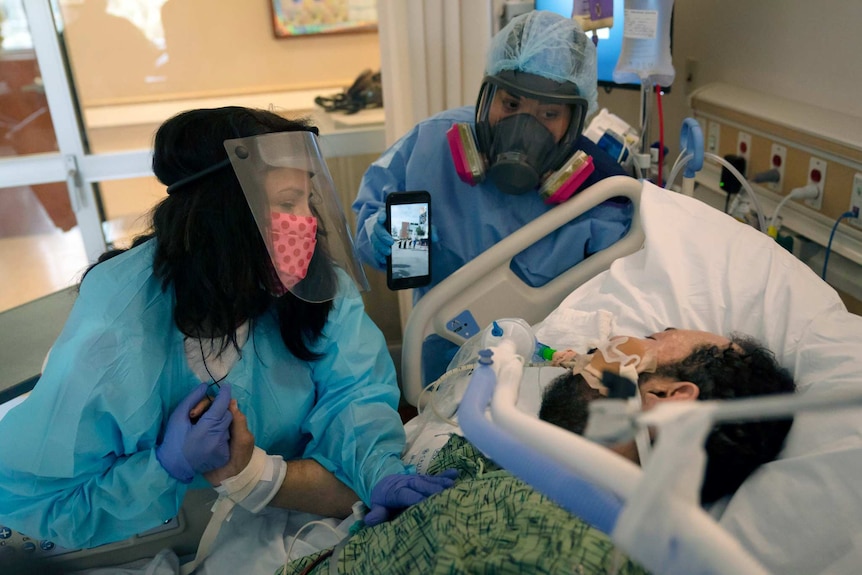 Two women in scrubs wearing face shields watch over a man with tubes coming out of his mouth on a bed