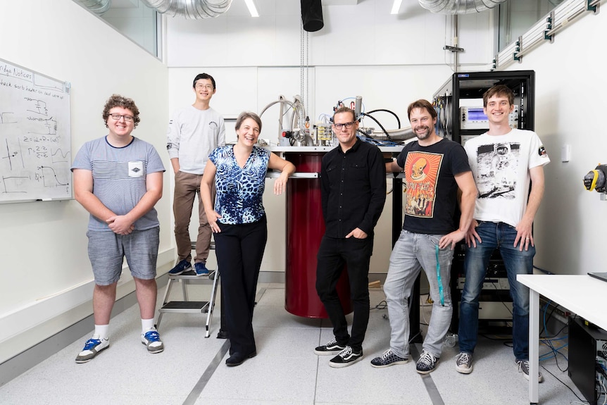 Six scientists stand casually and smile, with pipes and apparatus on a desk behind them.