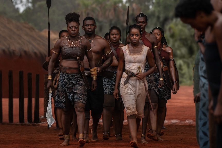 A group of Black, African warriors march through a a village in tribal battle gear. 